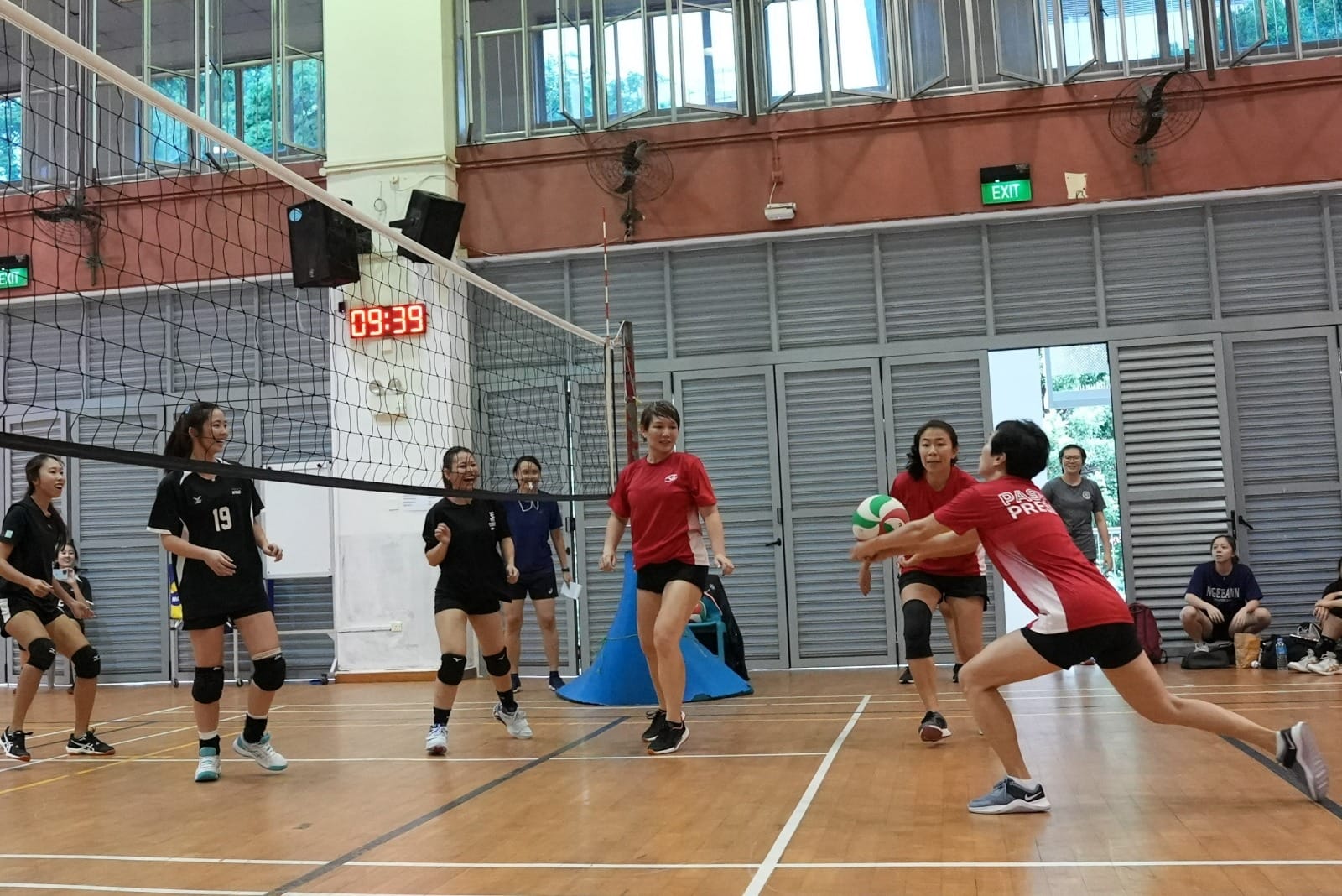Women in red and black uniforms play indoor volleyball, with a player in red bumping the ball near the net.